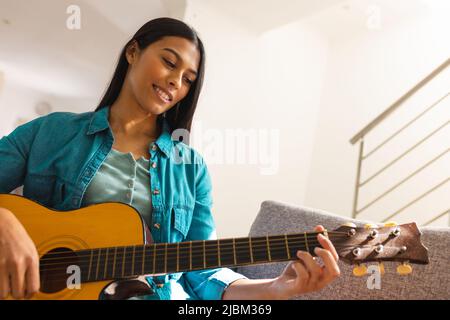 Jeune femme biracial souriante pratiquant la guitare à la maison Banque D'Images