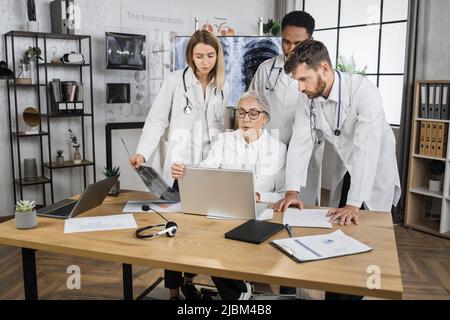 Équipe internationale de trois stagiaires ayant une réunion urgente avec une femme médecin senior dans la salle de conférence de l'hôpital. Blouses de laboratoire blanches utilisant un ordinateur portable moderne pour l'analyse du dossier médical des patients. Banque D'Images