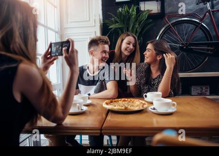 Jeune femme photographiant ses amis positifs dans une maison de café à l'aide d'un smartphone, arrière-plan défoqué. Banque D'Images