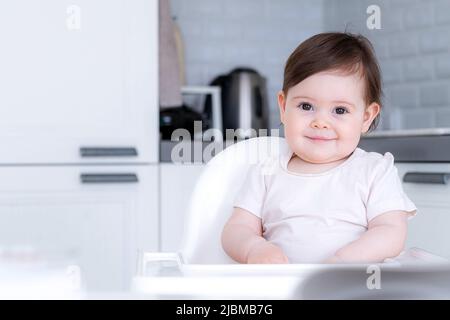 Portrait mignon bébé fille sur la cuisine assis sur une chaise haute, sourire enfant heureux de manger la première nourriture solide Banque D'Images