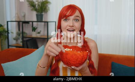 POV de la femme enthousiaste à tête rouge assis sur un canapé en mangeant du pop-corn et en regardant des séries, films, films ou contenus de médias sociaux intéressants à la maison. Ginger Girl appréciant les divertissements domestiques Banque D'Images