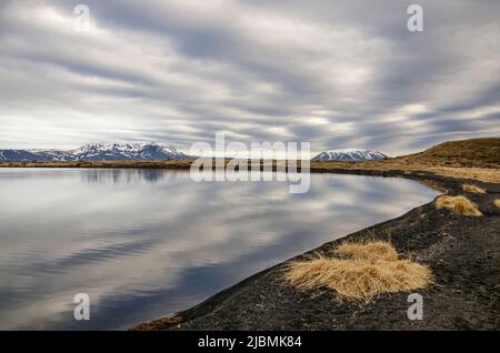 Vue depuis une plage de sable noir et de gravier sur un lac réfléchissant dans la région de Myvatn en Islande vers une chaîne de montagnes enneigée sous un drame Banque D'Images