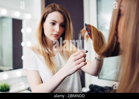 Jeune joli coiffeur portant des vêtements décontractés tenant une mèche de cheveux longs entre les doigts et coupant les extrémités dans le studio de beauté. Banque D'Images