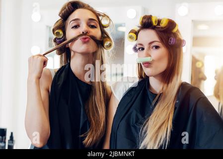 Deux jolies petites amies en cape noire portant des curlers faisant de la moustache de leurs cheveux et de rouleau dans le salon de coiffure. Banque D'Images