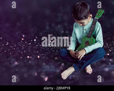 latino garçon assis sur le sol avec un ukulele dans ses bras riant et regardant vers le bas. Photo studio avec arrière-plan noir et espace de copie. Concept de le Banque D'Images