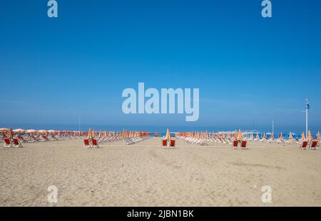 Une vue sur les chaises de plage rouges avec parasols orange et blancs par une journée ensoleillée avec un ciel bleu clair. Banque D'Images