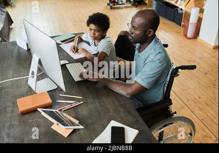 Père africain handicapé assis en fauteuil roulant à l'aide d'un ordinateur à table pour aider son fils à faire ses devoirs Banque D'Images