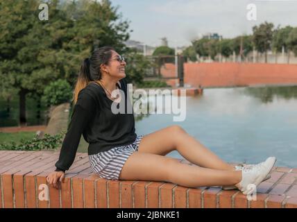 Une femme latine souriante et insouciante portant des lunettes de soleil est assise sur une place avec un étang tout en contempler le paysage urbain Banque D'Images