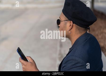 Latino gay mâle avec maquillage porter un chapeau tendance tenant un téléphone portable dans un parc Banque D'Images