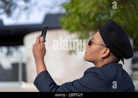 Latino gay homme avec maquillage sur le port de chapeau tendance regardant le téléphone cellulaire dans un parc Banque D'Images