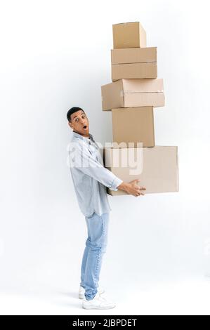 Surpassé homme de course mixte dans des vêtements décontractés élégants, livreur, debout sur fond blanc isolé, tenant de nombreuses boîtes en carton, livrant des colis, regardant dans l'étonnement à l'appareil photo, livraison Banque D'Images