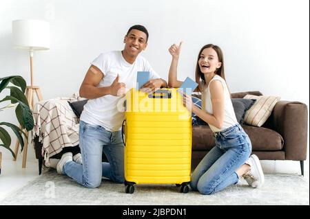 Joyeux jeune couple multiracial, un type hispanique et une fille caucasienne, assis sur le sol à la maison dans le salon près d'une grande valise jaune, a reçu un vaccin, en voyage après la quarantaine, souriant Banque D'Images