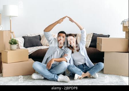 Joyeux heureux couple multiracial jeune dans l'amour assis sur le sol dans le salon de leur nouvelle maison entre des boîtes en carton, faisant un toit imaginaire avec leurs mains, regardant l'appareil photo, souriant Banque D'Images