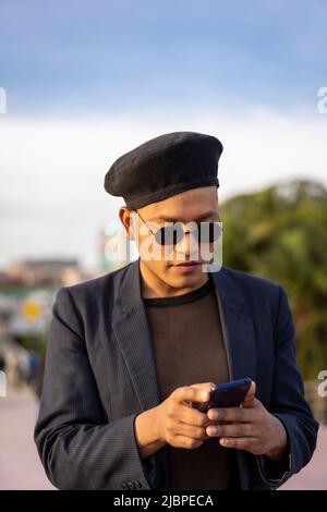 Latino gay homme avec maquillage sur le port de chapeau tendance regardant le téléphone cellulaire dans un parc Banque D'Images