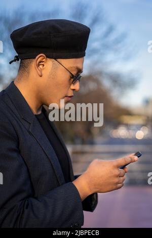 Latino gay homme avec maquillage sur le port de chapeau tendance regardant le téléphone cellulaire dans un parc Banque D'Images