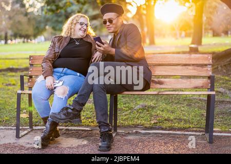 Amis gays assis sur un banc dans un parc au coucher du soleil bavardant et regardant le téléphone cellulaire Banque D'Images