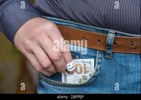 La main d'un homme tire cent dollars de sa poche de pantalon. L'index est enveloppé de ruban blanc. Un visage souriant est dessiné avec un marqueur noir Banque D'Images