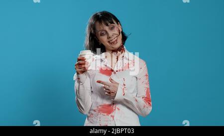 Un zombie à l'aspect inepte avec des cicatrices profondes qui sentent le café d'une tasse sur fond bleu. Monstre effrayant à l'aspect fou avec des blessures sanglantes ayant une tasse de café tout en grognant à la caméra. Banque D'Images