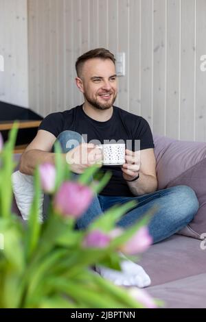 Un jeune homme souriant et souriant, en T-shirt noir et Jean, est assis sur un canapé confortable et dégusté une tasse de thé chaud. Repos et détente. Des moments heureux. Positif Banque D'Images