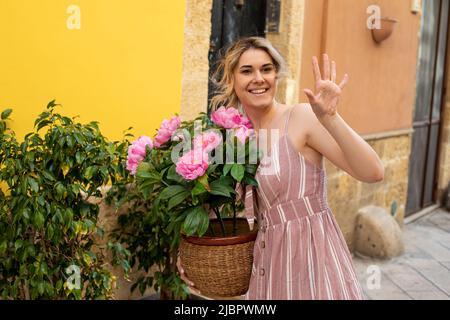 Jeune femme souriante portant une robe rayée rose d'été debout près des oliviers, tenant pot avec des pivoines roses, saluez. Banque D'Images