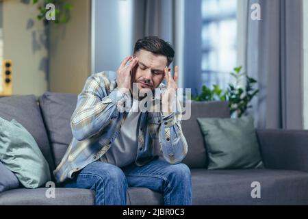 L'homme malade est assis seul à la maison sur le canapé, a un mal de tête sévère Banque D'Images
