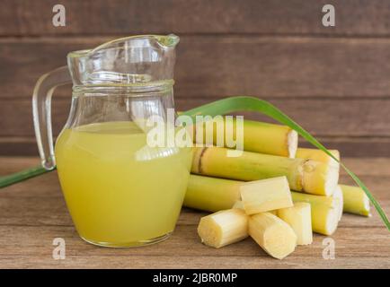 Jus de canne à sucre fraîchement pressé dans un pichet avec morceaux de canne coupés sur fond de bois. Banque D'Images