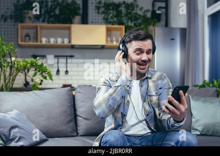 Un homme heureux à la maison souriant et écoutant de la musique à partir du téléphone utilise l'application de musique sur smartphone en ligne, avec de grands écouteurs Banque D'Images