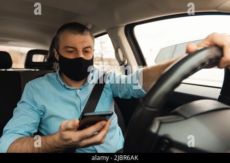 Homme conduisant une voiture en utilisant un smartphone portant un masque facial en automobile Banque D'Images