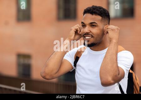 Jeune afro-américain portant des écouteurs sans fil, écoutant de la musique tout en marchant dans la ville, espace libre Banque D'Images