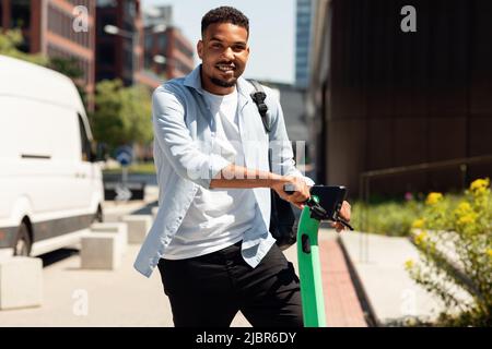 Concept de transport écologique. Heureux noir homme debout en face de la ville, tenant e-scooter et souriant à la caméra Banque D'Images