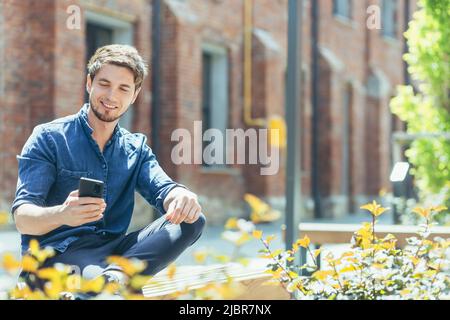 Jeune homme d'affaires devant le bureau assis sur un banc appréciant le téléphone souriant et heureux Banque D'Images