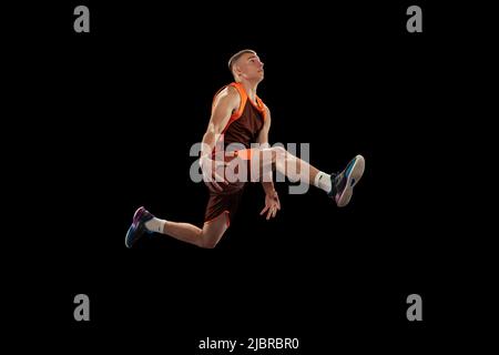 Portrait dynamique du jeune joueur professionnel de basket-ball en mouvement, sautant avec une balle isolée sur fond noir studio Banque D'Images