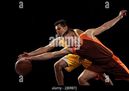 Portrait de deux hommes concentrés, joueurs de basket-ball en mouvement, dribbling isolé sur fond noir studio Banque D'Images