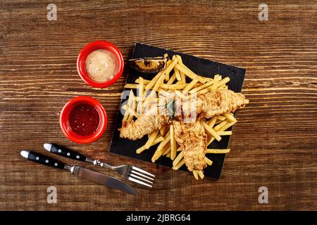 Assiette noire avec poisson et chips, mayonnaise et ketchup sur fond de bois Banque D'Images