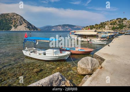 Petits bateaux amarrés dans un lagon bleu de mer Égée avec des îles montagneuses autour, plage en arrière-plan et drapeau turc en haut de la colline à Turunc, Mugla, Tu Banque D'Images