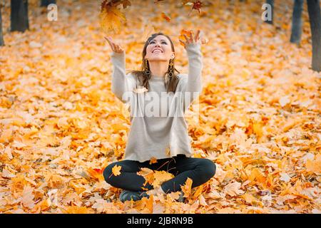 Une jeune femme souriante s'amusant dans un parc d'automne jette des feuilles mortes Banque D'Images