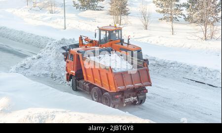 Un gros tracteur orange nettoie la neige de la route et la charge dans le camion.Nettoyage et nettoyage des routes de la ville de la neige en hiver Banque D'Images