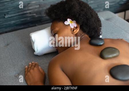 Femme afro-américaine avec les yeux fermés recevant un massage aux pierres chaudes à la maison Banque D'Images