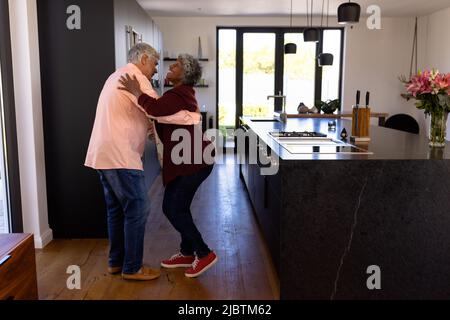 Vue latérale d'un couple senior romantique multiracial heureux dansant dans la cuisine à la maison de retraite Banque D'Images