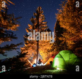 Silhouette d'homme assis près d'un arbre au milieu du camping et regardant un feu de camp lumineux. Bonne réacation active dans le camp le soir d'été sous ciel étoilé. Banque D'Images