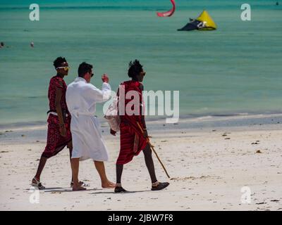 Paje, Zanzibar - janvier 2021: Guerriers africains de Maasai en costume traditionnel et homme caucasien dans un peignoir blanc marchant le long d'une plage de sable contre Banque D'Images