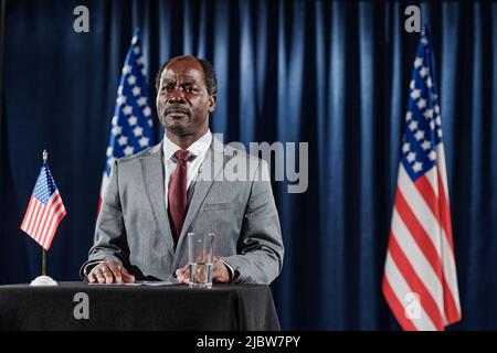 Homme politique afro-américain en costume debout à Tribune et présentant son rapport avec des drapeaux américains derrière lui Banque D'Images