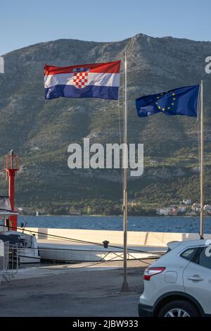 Drapeaux européens et croates à vent sur le quai de Korcula, en Croatie, avec Orebic dans la péninsule de Peljesac en arrière-plan Banque D'Images