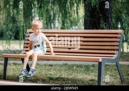 Un petit enfant joue dans le parc, monte sur un banc.Kid est assis sur un banc de parc. Concept de famille et d'enfance heureux. Bébé en bonne santé à l'extérieur. Grand arbre fol Banque D'Images