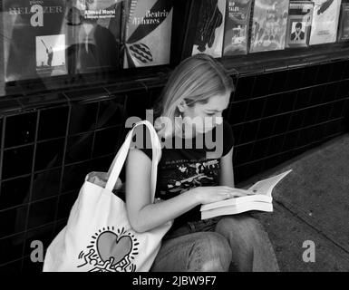 Une jeune femme lit un livre que vous venez d'acheter sur le trottoir après avoir visité la célèbre librairie City Lights Booksellers à San Francisco, en Californie. Banque D'Images