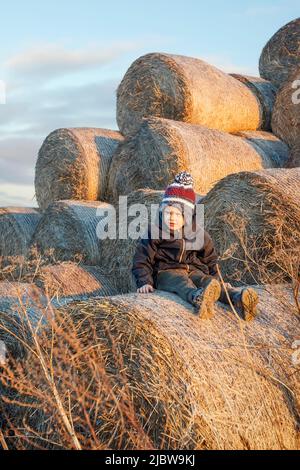 Un joli petit garçon en vêtements d'automne et un bonnet tricoté se pose sur un fond de balles de foin dorées. Photo verticale. Banque D'Images