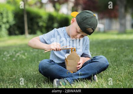 Enfant mangeant des nouilles udon assis sur l'herbe. Sale tache de sauce sur son t-shirt. Nouilles qui sortent de votre bouche. À l'extérieur Banque D'Images