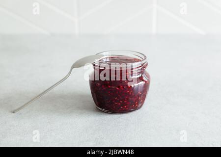 Confiture de framboises rouges maison naturelle dans un petit pot en verre avec cuillère sur fond gris. Vue avant, mise au point sélective. Banque D'Images