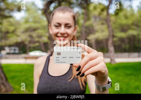 Bonne femme brune portant un soutien-gorge de sport noir debout sur le parc de la ville, à l'extérieur montrant une carte de crédit ou de débit en plastique, effectuant des paiements en ligne. Sélectif Banque D'Images