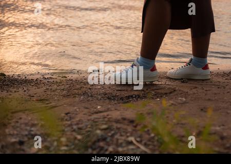 Femme en peau sombre et écourtée jambes en bottes marchant près de l'eau de la rivière ou côté mer sable, plage d'herbe, vacances nature à l'étranger Banque D'Images